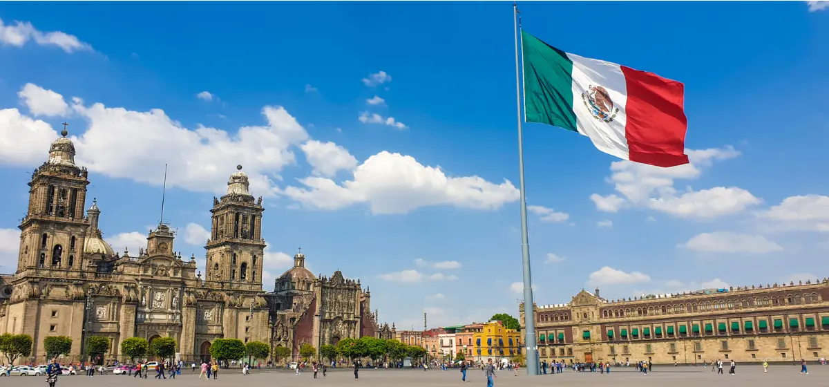 Una gran bandera mexicana ondea en la plaza del Zócalo de la Ciudad de México, con la Catedral Metropolitana y el Palacio Nacional al fondo bajo un cielo azul.
